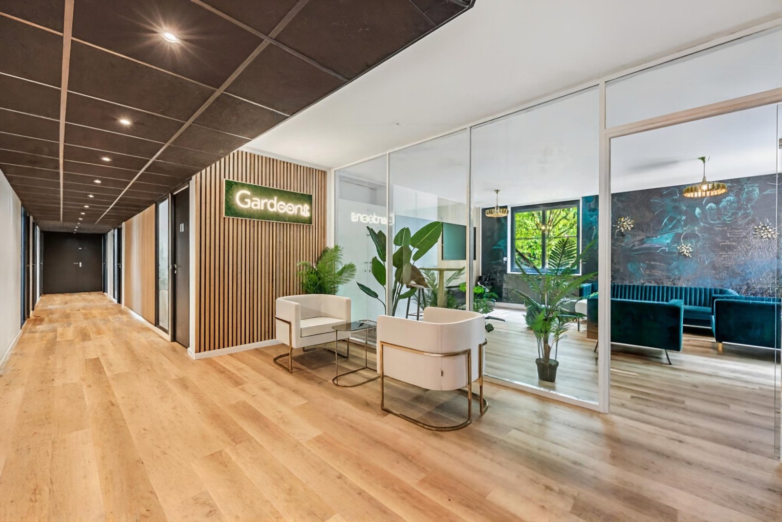 Accueil du Garden : enseigne lumineuse, mur en lames de bois, fauteuils blancs et plantes, salon vitré en arrière-plan.